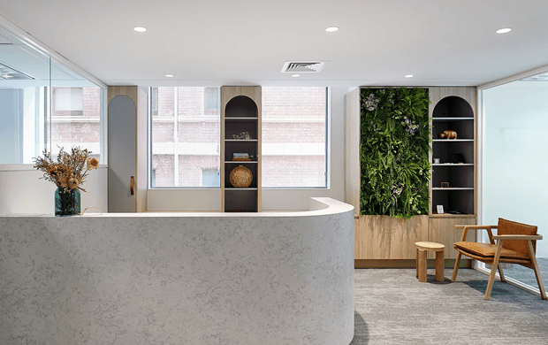 Contemporary reception or waiting area with a large curved light stone reception desk, integrated arched shelving, floor-to-ceiling windows, a vertical living plant wall, pale timber cabinetry, and mid-century-style lounge seating on a grey carpet.