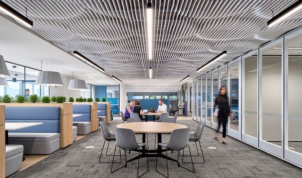 A bright contemporary commercial office interior with an open collaborative lounge and meeting area. The space features round meeting tables with upholstered chairs, booth seating with timber dividers, glass-fronted private offices, carpet tile flooring, and a distinctive undulating linear ceiling grid with integrated strip lighting. Large windows provide abundant daylight and views of an urban setting.