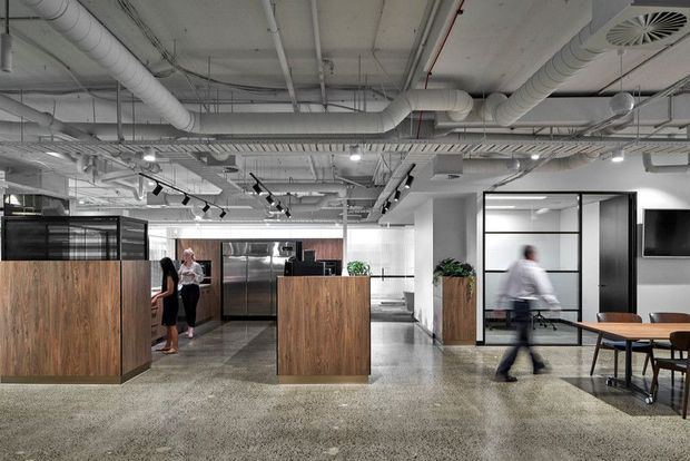 A modern open-plan commercial interior with exposed white services and ductwork, polished concrete flooring, timber-clad reception or service counters, black-framed glazed meeting rooms, track lighting, and a dining or collaboration table. The image has an industrial office fit-out with a neutral grey, white, black, and warm timber palette.