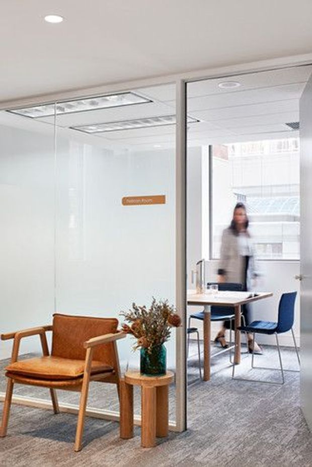 A bright contemporary commercial office interior viewed through a glass-partitioned meeting room. The foreground features a tan upholstered timber armchair and small round timber side table with a vase of dried foliage. Beyond the glazing is a compact meeting area with a light table, blue chairs, grey carpet and a blurred person. The space has white walls, a suspended acoustic ceiling with recessed lighting, extensive internal glazing and a small wall sign.