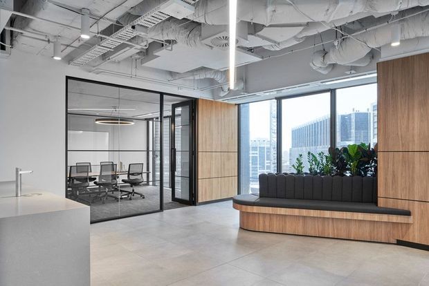 A bright contemporary commercial office interior with an exposed white services ceiling, polished concrete-look flooring, glass-partitioned meeting room, and a built-in timber-clad window bench with dark upholstered seating and integrated planting. Large windows overlook a dense urban skyline.