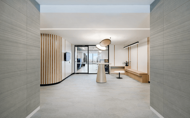 A refined contemporary commercial lobby or reception area with pale grey textured wall panels, light flooring, curved timber slat screening, glass partitioning, a sculptural central pendant light, linear ceiling lighting, and built-in timber bench seating. The space has a clean, calm, minimalist fit-out with subtle warm timber accents.