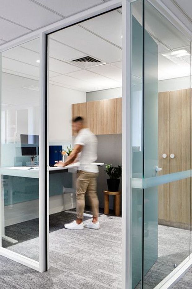 A bright contemporary commercial office interior viewed through extensive glazed partitions and a glass door. The space features grey carpet flooring, white acoustic ceiling tiles with recessed lighting and air-conditioning vents, pale blue-grey walls, light timber built-in cabinetry, white desks, and a compact workstation. A blurred person is standing at the desk, with plants and office equipment visible.