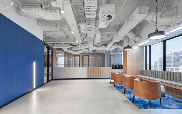 Contemporary commercial interior with an open lounge or breakout area, polished concrete flooring, exposed white services and ductwork, bold blue feature wall, timber screening, and floor-to-ceiling windows overlooking an urban setting. Brown upholstered lounge chairs, banquette seating, pendant lighting, and a blue patterned rug define the seating zone.