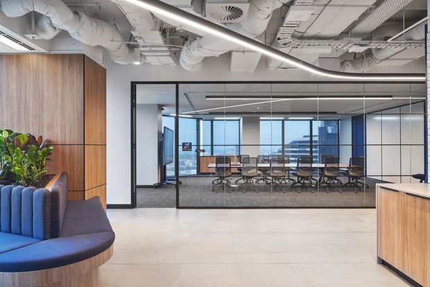 Contemporary commercial office interior with a large glazed meeting room, exposed services ceiling, linear integrated lighting, timber wall panelling and reception joinery. A curved upholstered banquette with planting occupies the foreground, while the meeting room contains a long conference table and office chairs with city views beyond.