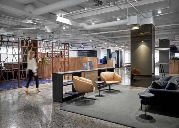 Open-plan contemporary commercial interior with a lounge and informal meeting area. The space features polished concrete flooring, exposed industrial-style services and ductwork, suspended linear lighting, dark built-in partitions, timber and metal screening, and a mix of tan lounge chairs and a dark sofa arranged on a large rug.