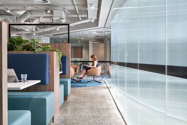 A contemporary open-plan office interior with a polished concrete floor, exposed white services and ductwork, glass partitions, timber privacy screens, integrated blue upholstered booth seating, indoor planting, and casual lounge chairs. The space has abundant daylight and a collaborative commercial workplace atmosphere.