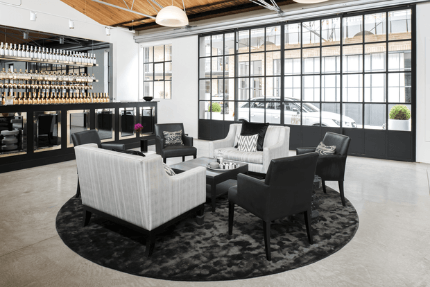 A refined commercial lounge and reception area with a seating arrangement on a round dark rug, surrounded by large black-framed industrial-style windows. A built-in bar or display wall with extensive bottle shelving occupies the left side. The space features polished concrete flooring, white walls, exposed timber ceiling panels with visible services, and pendant lighting.
