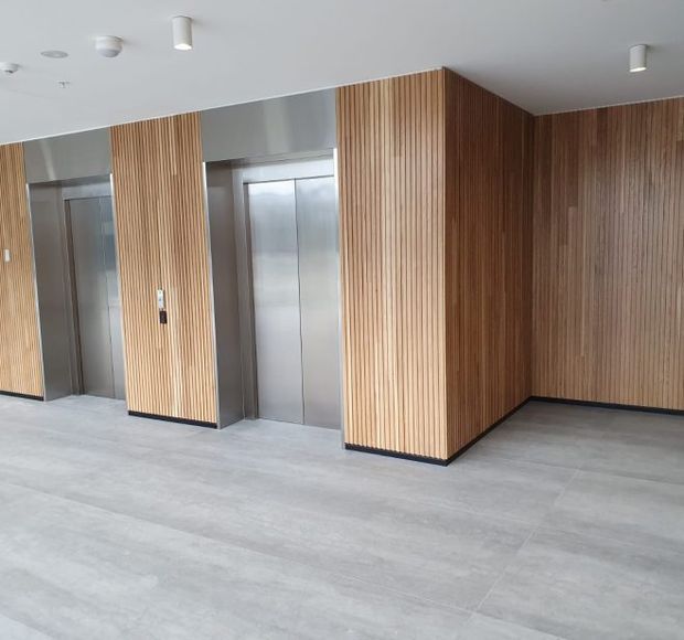 Modern elevator lobby with floor-to-ceiling vertical timber slat wall panelling, stainless steel elevator doors and surrounds, a light grey tiled floor, and a plain white ceiling with recessed and surface-mounted lighting. The space is clean, minimal, and likely part of a commercial or multi-storey building.
