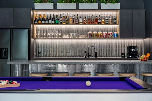 A sophisticated home bar with a full-size purple-felt pool table in the foreground. The bar features dark built-in cabinetry, illuminated open liquor and glassware shelving, a ribbed metallic backsplash and bar front, a stone-look countertop, under-cabinet lighting, a sink, coffee machine and refrigerator. Planters sit above the shelving, adding greenery to the otherwise dark, polished interior.