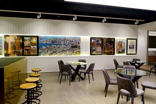 A contemporary commercial café or hospitality interior with polished concrete flooring, a ribbed white feature wall displaying framed artwork and graphics, black ceiling track lighting, and multiple small dining tables with upholstered chairs. A curved bar counter with stools occupies the left side, while a service or kitchen area is partially visible at the rear right.