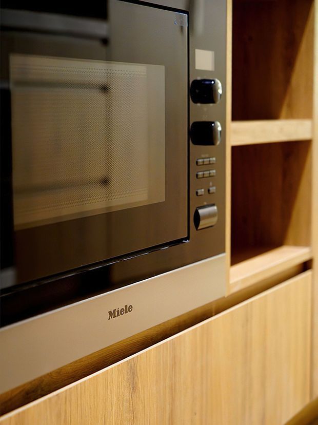Close-up of a built-in Miele microwave or oven integrated into warm-toned timber cabinetry, with open display shelving visible to the right. The cabinetry has a natural light wood finish and the appliance has a dark reflective glass front with metallic controls.