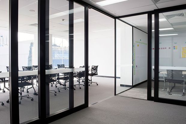 A bright contemporary office interior with black-framed glass partition walls and doors creating enclosed meeting rooms or workspaces. White ceilings with recessed fluorescent lighting, white walls, grey carpet, and minimal office tables with black wheeled chairs are visible. Collaborative planning notes and boards appear inside the glazed rooms.