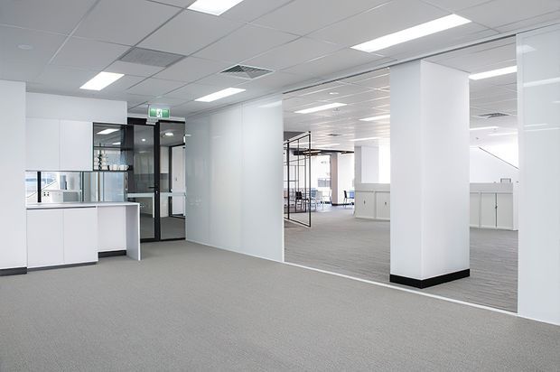 Bright, open-plan commercial office interior with white partition walls, glazed meeting-room doors, exposed suspended ceiling tiles, recessed fluorescent lighting, grey carpet, white built-in cabinetry, and structural columns. The space appears contemporary and minimally furnished.