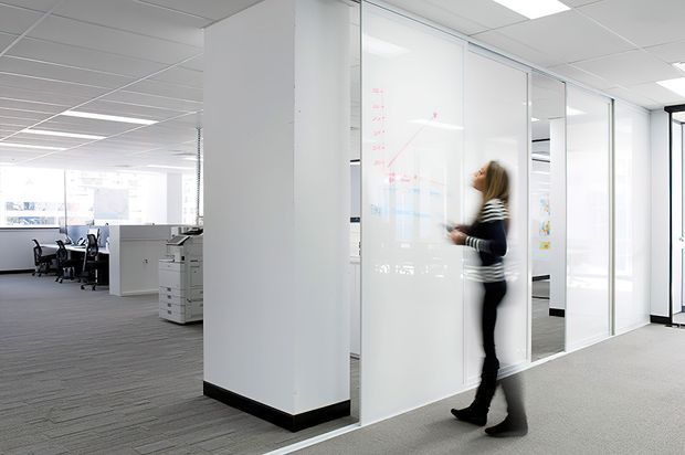 Bright contemporary open-plan office interior with grey carpet, white walls and ceiling, glass-partitioned meeting or work rooms, office desks, chairs, and a copier. A person is walking beside a translucent glass writing surface.