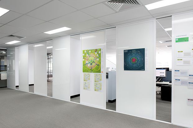 A bright modern commercial office interior with a carpeted open-plan workspace enclosed by white framed glass and solid partition panels. Several mounted maps and information graphics decorate the partitions, while desks and computer workstations are visible through the glazing. The space has a suspended acoustic ceiling with recessed fluorescent lighting and air-conditioning vents.