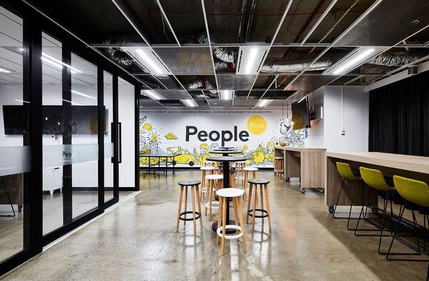 Contemporary open-plan commercial interior with polished concrete flooring, exposed services and ductwork, linear pendant lighting, black-framed glazed meeting rooms, and a collaborative breakout area furnished with stools and high tables. A large illustrated feature wall reading “People” adds a playful branded focal point, while a long timber-toned communal counter with lime-green stools lines the right side.