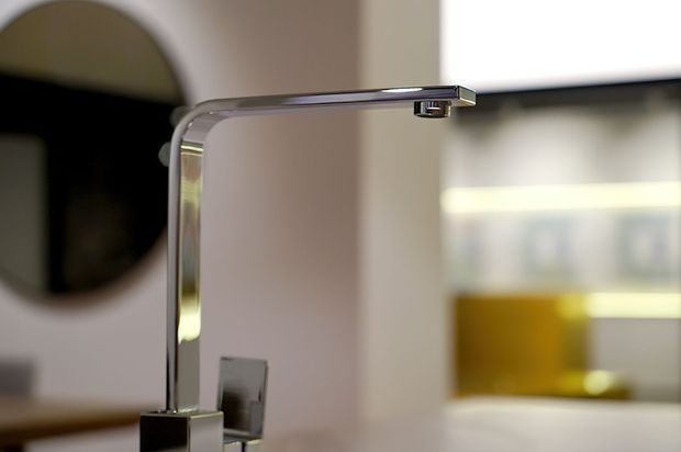 Close-up of a contemporary chrome kitchen faucet with a squared vertical body and flat horizontal spout. The faucet is mounted on a light-coloured benchtop, with softly blurred modern cabinetry or shelving in the background and a dark circular wall feature at left.