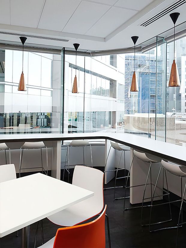 Bright contemporary commercial interior with a communal breakout or dining area beside full-height glazing. White angular tables, curved white counters, numerous high stools, and distinctive copper pendant lights create a clean, modern workplace hospitality setting. Dark flooring contrasts with the predominantly white fit-out, while an urban building exterior is visible through the windows.