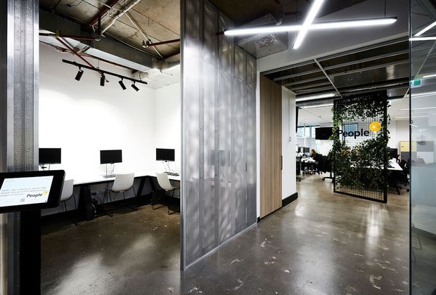 Contemporary open-plan commercial office interior with exposed services, polished concrete floors, translucent corrugated metal wall panels, glass partitions, linear LED lighting, and a planted screen separating a workstation area from a meeting or collaboration space. The palette is predominantly white, grey, black, and concrete tones.