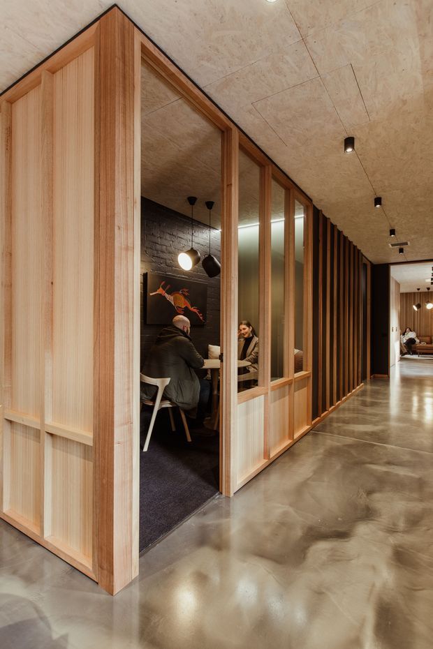 Interior view of a contemporary commercial office or hospitality workspace, featuring a long timber-framed glazed partition with vertical timber battens, an enclosed meeting booth, dark exposed brickwork, and a polished concrete floor. The warm timber screening contrasts with black ceiling detailing and discreet recessed and pendant lighting. Additional seating and work areas are visible down the corridor.
