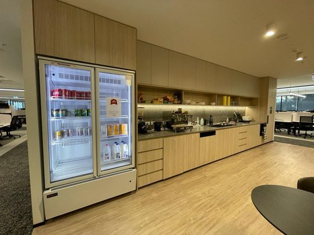 A contemporary commercial office kitchen and breakout area with extensive light wood cabinetry, a long dark benchtop, open illuminated shelving, integrated appliances, and a tall glass-front drinks refrigerator. The space has light timber-look flooring, recessed ceiling lighting, and carpeted office work areas visible beyond the kitchen.