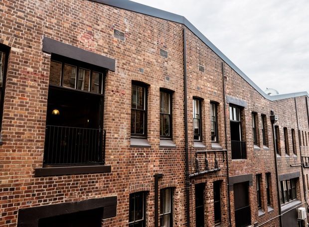 Exterior view of a multi-storey brick building with a long industrial-style facade, repeated tall windows, dark metal window and door frames, and a low-pitched roof edge. The weathered red-brown brickwork and regular openings create a warehouse or converted industrial character.