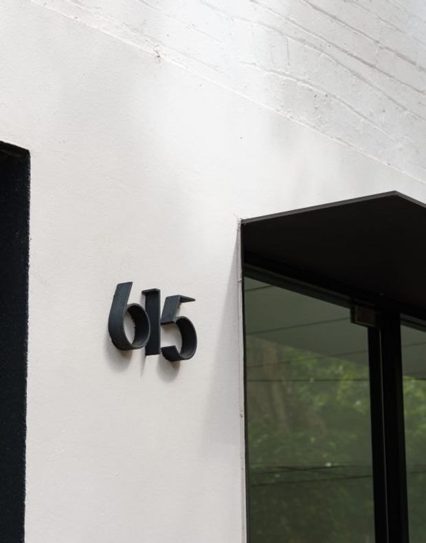 Close-up of a contemporary building entrance showing a white rendered wall with black raised house numbers “65” beside a black-framed glazed doorway beneath a dark projecting canopy. The image focuses on the facade and entry detailing.