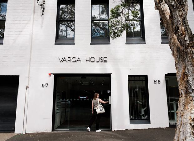 Street-level photograph of the white-painted brick facade of Varga House, with tall black-framed windows, a glazed commercial entrance, street numbers, signage, and a mature tree in the foreground.