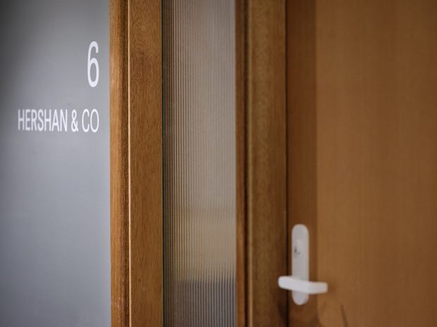 Close-up architectural detail of a timber-framed door with a vertically ribbed translucent glass panel and white lever hardware. A grey wall beside the doorway is marked with the number 6 and the text “HERSHAN & CO,” suggesting an entry to a commercial or office space.