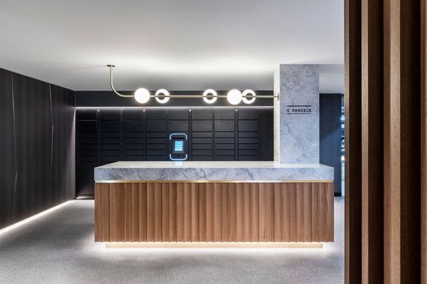 A sophisticated commercial reception area featuring a sculptural timber reception desk with vertical fluting, a pale stone benchtop, dark wall panelling, integrated linear lighting, and a contemporary circular pendant fixture. A parcel collection point is visible behind the desk, and the space is framed by dark timber elements.