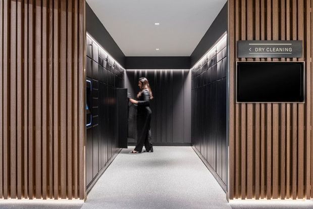 A sleek, dark-toned commercial interior resembling a hotel or apartment building service lobby. A central carpeted corridor is flanked by black built-in lockers or cabinetry with integrated illuminated shelving. Vertical timber slat screens frame both sides of the entrance, while a wall-mounted sign reads “DRY CLEANING” above a black display or service panel. A person stands near the lockers.