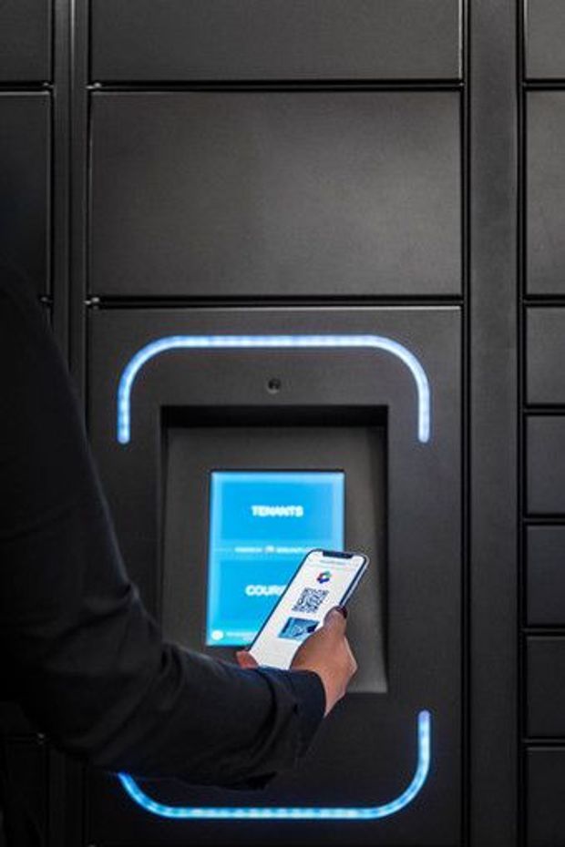 Close-up of a dark, built-in locker or access-control panel with a glowing blue illuminated outline around a recessed touchscreen. A person is holding a smartphone displaying a QR code toward the screen, suggesting contactless access or locker pickup technology. The surrounding surface is matte black and panelled with integrated seams.