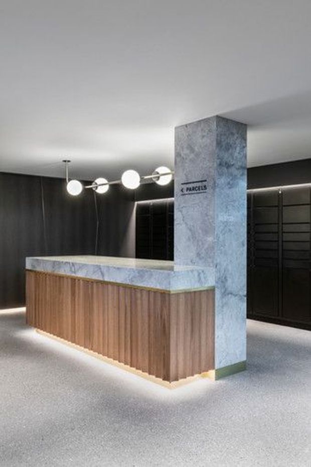 A contemporary commercial reception or concierge area featuring a wood-slatted desk with a pale stone benchtop, a tall textured stone-clad column, dark wall panelling and cabinetry, illuminated perimeter detailing, and spherical pendant lighting.