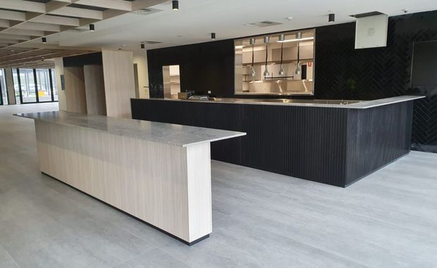 A spacious contemporary commercial kitchen or hospitality fit-out with a large light-wood island in the foreground and a dark fluted island counter behind it. The space features a pale concrete-look floor, black feature walls, timber-look cabinetry, a service or display opening with shelving, and a coffered ceiling with recessed and pendant-style lighting.