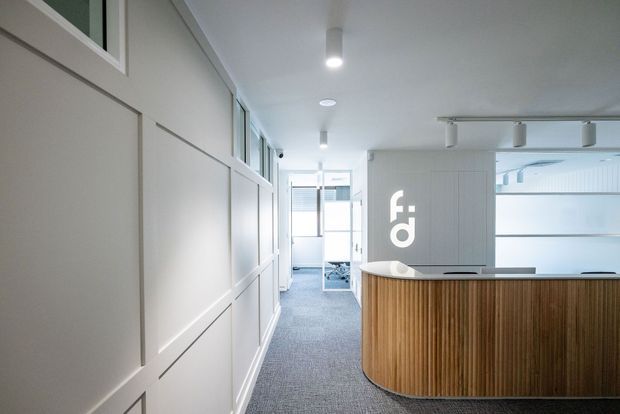 Bright contemporary commercial reception area with a curved ribbed timber reception desk, white paneled wall detailing, grey carpet, recessed and track lighting, and glazed office spaces beyond.