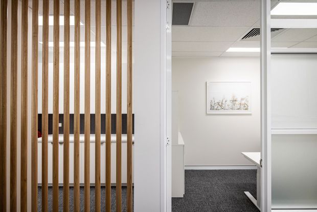 A contemporary commercial office interior viewed through a glazed partition and vertical timber screening. The space features white walls, grey carpet, suspended acoustic ceiling tiles with recessed lighting and air-conditioning vents, built-in white cabinetry, and a minimalist framed artwork. A partially visible workstation is seen on the right.