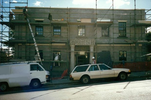 Street-level view of a multi-storey historic building undergoing exterior restoration or construction. Scaffolding covers the facade, with workers, ladders, temporary fencing, and parked utility vehicles in front. The building has symmetrical window bays and an ornate central entrance with columns and decorative stonework.