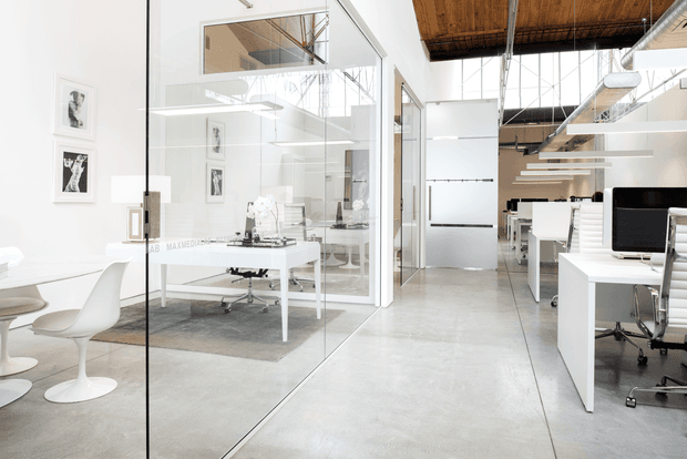 Bright contemporary open-plan office interior with glass-partitioned meeting rooms, white desks and ergonomic office chairs, polished concrete flooring, exposed timber ceiling elements, exposed services, and linear suspended lighting. A small informal seating area is visible at left.