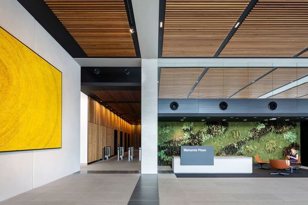 A modern commercial lobby or reception area with a large white reception desk, a green living wall, and visible “Manunda Place” signage. The space features a dramatic yellow wall artwork, large-format grey floor tiles, timber slatted ceiling panels with integrated lighting, exposed dark structural elements, and a small seating area. The composition emphasizes a spacious, refined corporate interior with biophilic planting and strong geometric lines.