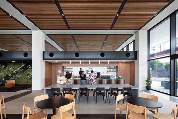 A contemporary café or hospitality space with a prominently framed service counter and seating area. The interior features a warm slatted timber ceiling, timber wall panelling behind the counter, black metal framing and ducts, large floor-to-ceiling windows, communal and round café tables, and light timber chairs. A planted feature wall and outdoor urban views are visible at the edges of the image.