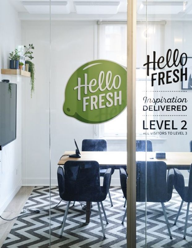 Bright contemporary office meeting room viewed through a glazed partition, with a light timber-framed glass wall, white walls and ceiling, a meeting table with dark upholstered chairs, and a bold black-and-white geometric carpet. Green and black window decals display “Hello Fresh” branding and visitor information. A wall-mounted screen and small plant shelves are visible in the adjacent area.
