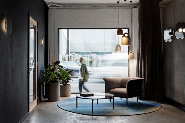 A dark, refined commercial-style lounge or reception space with charcoal textured walls, exposed ceiling services, pendant lighting, and a large street-facing window. A sculptural armchair and round coffee table sit on a blue circular rug, accompanied by a large potted plant and framed wall artwork. The urban streetscape is visible through the glazing, with a blurred passerby outside.
