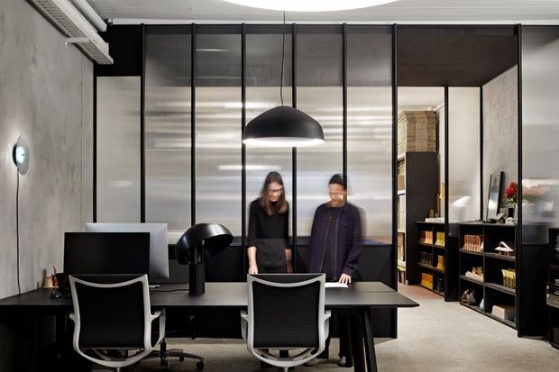 A sophisticated contemporary office interior with a long dark workstation and two ergonomic task chairs in the foreground. Black-framed glazed partitions divide the workspace, while dark built-in shelving and storage line the right side. Concrete-look walls, a pale carpeted floor, black pendant lighting, and a large circular ceiling feature create a refined, high-contrast commercial environment.