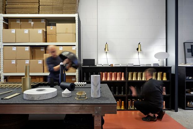 A contemporary industrial-style commercial workspace or design showroom with open shelving, stacked cardboard boxes, product samples, and display objects. A large dark worktable occupies the foreground, while black shelving in the background holds rows of colourful cylindrical items and materials. The space has white tiled walls, black metal framing, warm orange flooring, and task and display lighting. Two blurred people are working in the space.