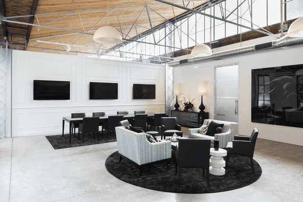 A spacious open-plan loft-style interior combining a living room and dining area. The room features polished concrete flooring, white paneled walls, expansive industrial steel-framed glazing, exposed timber ceiling boards, visible roof structure and ductwork, sculptural pendant lights, black rugs, and predominantly black-and-white contemporary furnishings. A large dark artwork or reflective panel is mounted on the right wall.