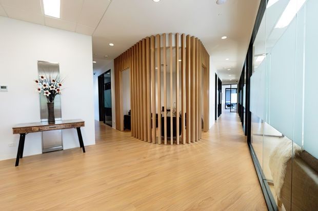 A bright contemporary commercial interior lobby or reception area with pale timber flooring, white walls and ceiling, extensive floor-to-ceiling glazing, and a distinctive curved cylindrical timber-slat screen enclosing a central workspace or reception zone. A slim console table and floral arrangement sit against the left wall, while a corridor extends into the background.
