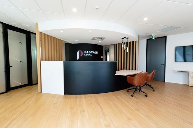 Bright contemporary commercial reception area with a curved black reception desk, white front panel, vertical timber slat feature walls, light timber flooring, black-framed glazed doors, and a small meeting/work table with rust-coloured office chairs. A branded wall sign is positioned behind the desk.