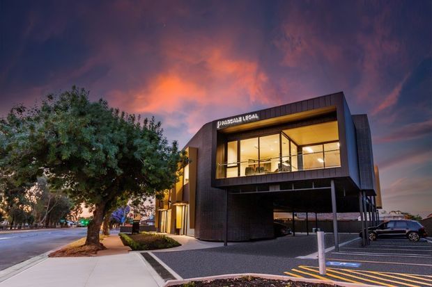 Twilight exterior photograph of a contemporary two-storey commercial building with a dark brick or masonry facade, projecting upper-level glazed office volume, illuminated interiors, covered ground-level parking, and landscaped street frontage. The building signage indicates a legal office.