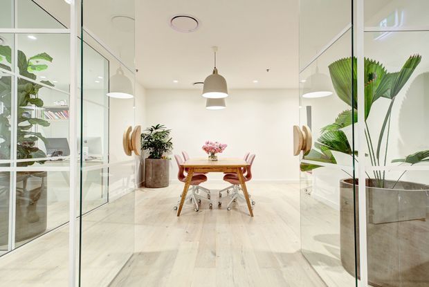Bright, minimalist meeting or dining room viewed through full-height glass partitions. The space features pale timber flooring, white walls and ceiling, a central timber table with pink upholstered chairs, simple pendant lighting, and large potted plants reflected in the glazing. The glass enclosure and mirrored reflections create a spacious, polished commercial-office feel.